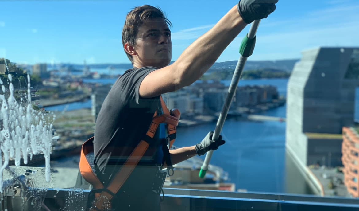 Oliver Olaussen i sele og sikringsutstyr mens han vasker glass høyt over Oslo, med byen og fjorden bak seg.